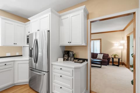 A modern kitchen with white cabinets and stainless steel refrigerator, opening to a bright living area.