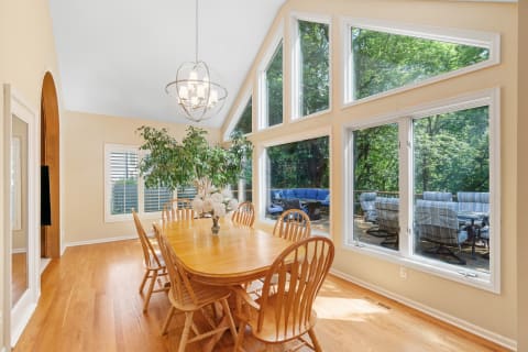 Light-filled dining room with wooden table and chairs, featuring large windows overlooking greenery.
