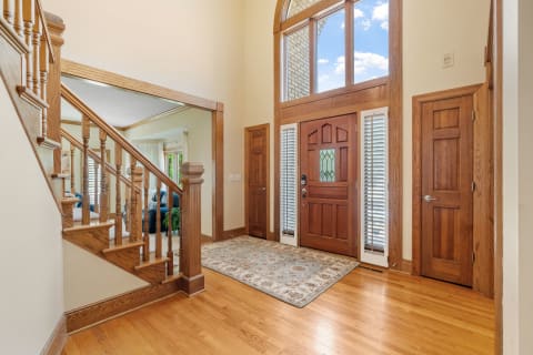 A spacious and elegant hallway with a staircase, wooden front door, and an area rug.