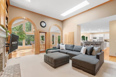 Spacious living room featuring a sectional sofa, stone fireplace, and views of a green outdoor space through large windows.