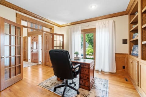 Interior view of a home office featuring a wooden desk, black chair, and large windows with curtains.
