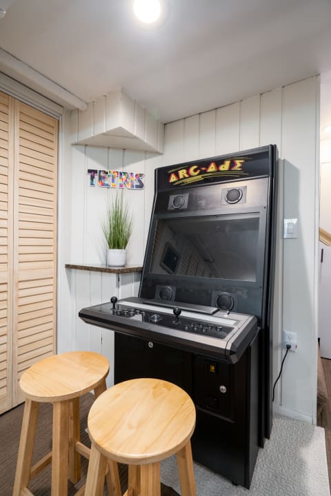 Arcade machine in a cozy room with wooden stools and a plant nearby.