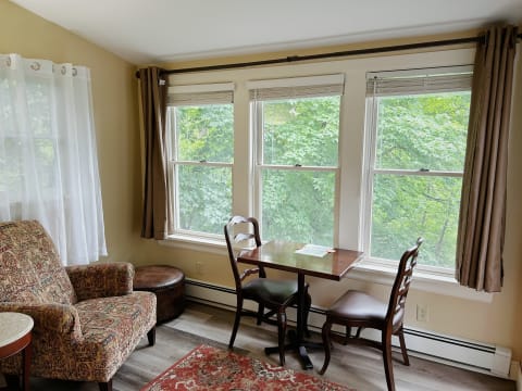 Cozy corner seating area featuring a patterned armchair, a small table with two wooden chairs, and bright windows overlooking greenery.
