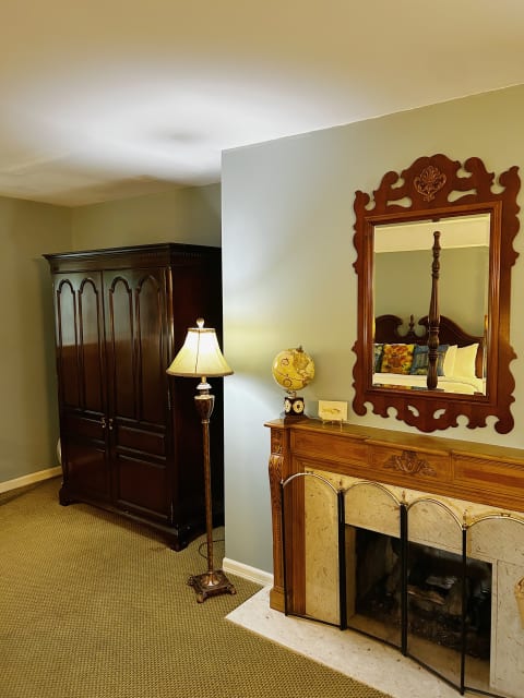 A traditional fireplace with a wooden mantle and an ornate mirror above it, complemented by a wooden armoire and a floor lamp in a cozy room.