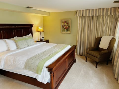 A spacious bedroom with a wooden bed, reading chair, and floral artwork, illuminated by soft light.