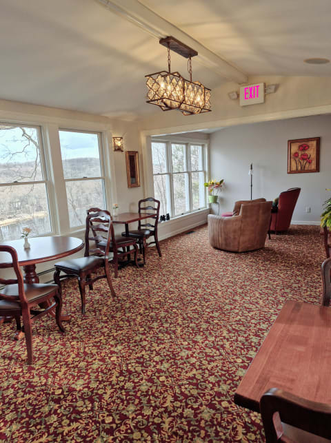 A bright communal area with wooden tables, chairs, a cozy armchair, and large windows offering scenic views.