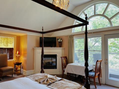 A sophisticated four-poster bedroom with a fireplace, arched windows, and a small dining area.