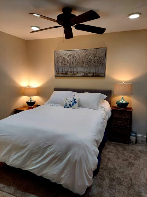 A serene bedroom featuring a queen-sized bed with white linens, warm lighting from bedside lamps, and a ceiling fan.