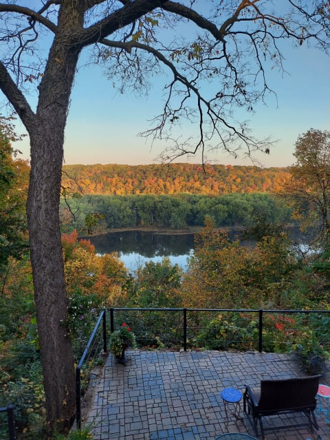 A peaceful scene with a river and colorful autumn trees, featuring a stone patio and a wicker chair.
