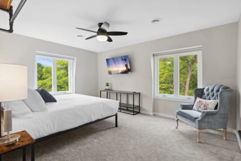 Bright modern bedroom with a large bed, two windows showing greenery, and a blue accent chair.