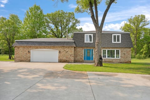 A picturesque two-story home with a blue door and surrounding green trees.