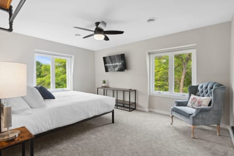 Modern bedroom with a large bed, armchair, and windows overlooking greenery.