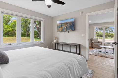 A spacious modern bedroom with a white bed, large windows showcasing trees outside, and a TV mounted on the wall.