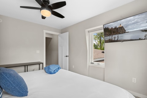 A modern bedroom featuring a white bed with blue pillows, a ceiling fan, and a mounted TV showing a lighthouse scene.