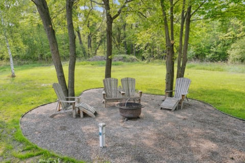 Adirondack chairs and fire pit in a grassy outdoor area surrounded by trees.