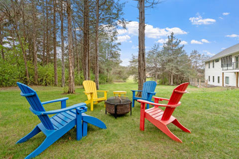 Colorful Adirondack chairs arranged around a fire pit in a green outdoor setting with trees and a house in the background.