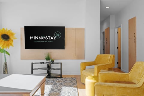 A living room with MINNēSTAY logo on TV, yellow chairs, and a sunflower.