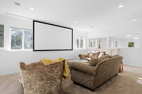 A cozy living room featuring a retractable projection screen with plush sofas and natural light from large windows.