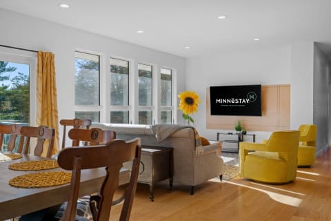A modern living room featuring a cozy couch, bright yellow chairs, a wooden dining table, and large windows with a view.