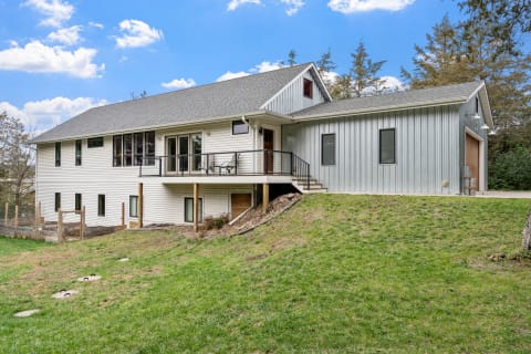 A modern home featuring a mix of white siding and gray metal panels, situated on a green slope under a clear blue sky.