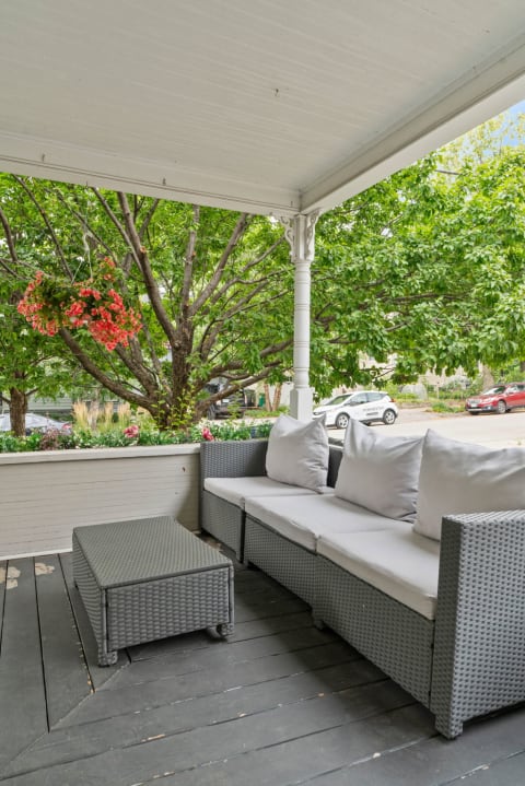 A cozy outdoor seating area with a gray wicker sofa, white cushions, and vibrant green foliage.