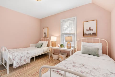 Children's bedroom featuring two white metal beds, pastel pink walls, and soft floral bedding.