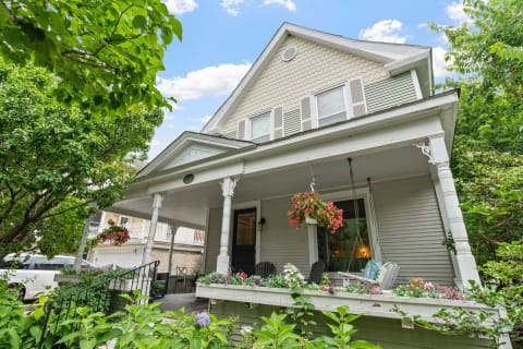 A two-story gray house with a flower-filled porch and lush greenery surrounding it.