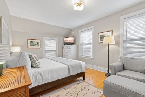 A cozy bedroom featuring a queen-sized bed, a nightstand, a modern dresser, and an armchair, illuminated by natural light.