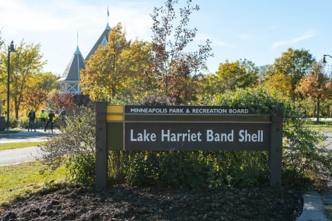 Sign for Lake Harriet Band Shell with autumn background and cyclists in the park.