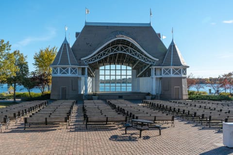 A view of an outdoor performance stage with benches set up facing it, surrounded by autumn trees and a body of water in the background.