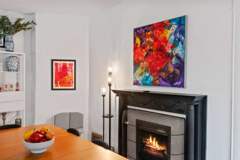 Modern dining space with a wooden table, vibrant fruit bowl, abstract art, and a lit fireplace.