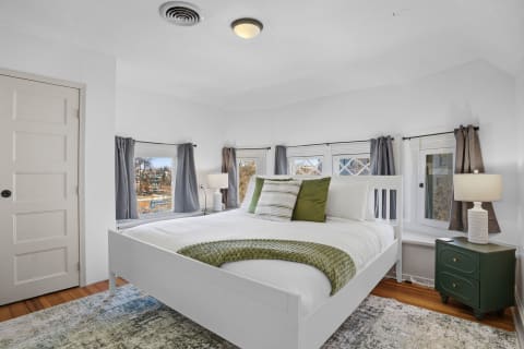 A bright corner bedroom featuring a white bed with green pillows and a bedside table with a lamp, illuminated by sunlight.
