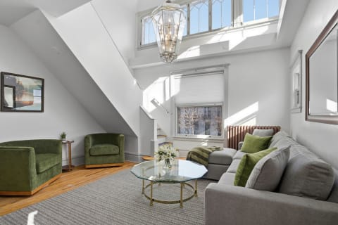 Bright living room with gray sectional sofa, green chairs, and glass coffee table under a skylight.