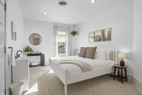 Bright bedroom with a white bed, potted plant, and cityscape artwork.