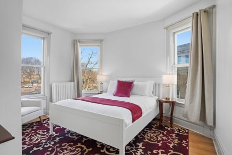 A cozy bedroom with a white bed, purple accents, and tall windows showcasing a city view.