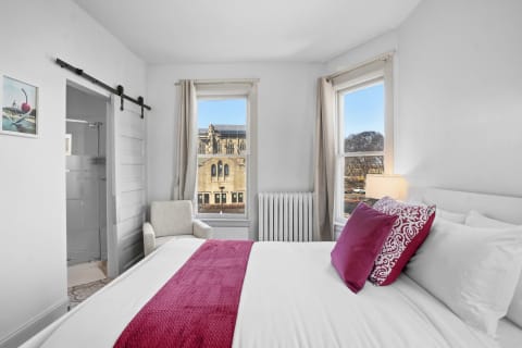 A modern bedroom showcasing white linens, vibrant pillows, and a view of a historic building through large windows.