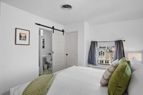 A cozy bedroom featuring white linens, green throw pillows, and a view of a historic building outside the window.