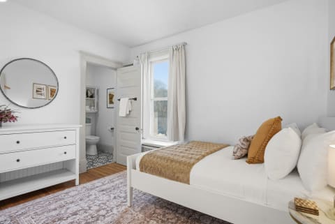A cozy bedroom with white walls, a neatly made bed, a dresser, and an open doorway leading to a bathroom.