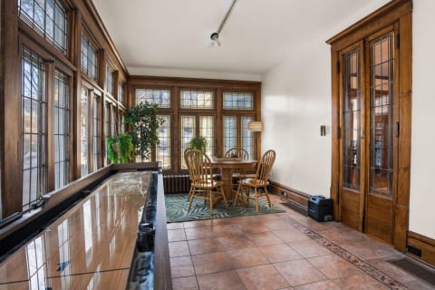 A sunlit porch with wooden details, a dining table, and large windows filled with greenery.