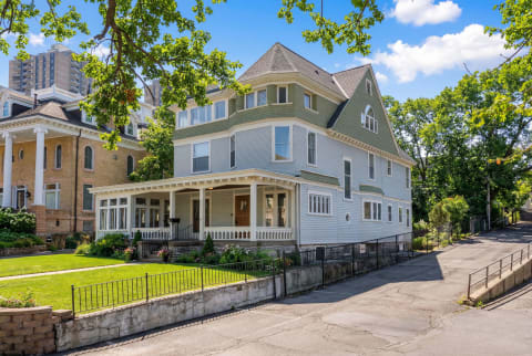 A Victorian-style house with a front porch and manicured lawn, flanked by other historic and modern buildings.