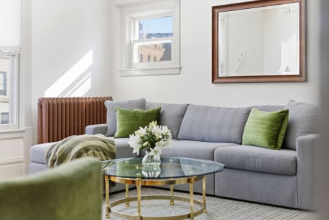 A stylish living room with a grey sectional sofa, green pillows, a glass coffee table, and a vintage radiator.