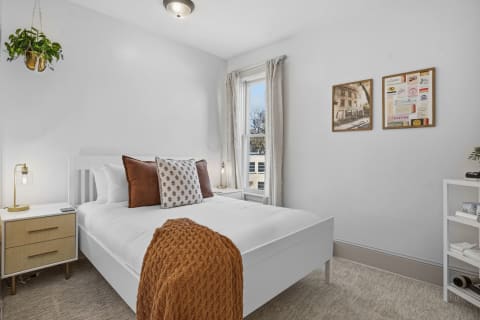 Bright and airy bedroom featuring a white bed with decorative pillows, a nightstand with a lamp, and framed art on the walls.