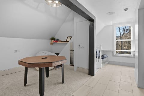 A bright upstairs room with a game table, chairs, and a window showcasing a view of trees.