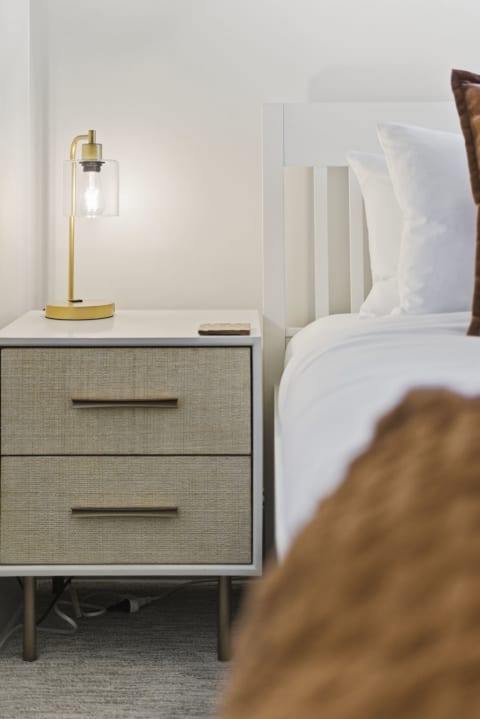 A cozy bedroom with a modern nightstand and bedside lamp next to crisp white linens on a bed.