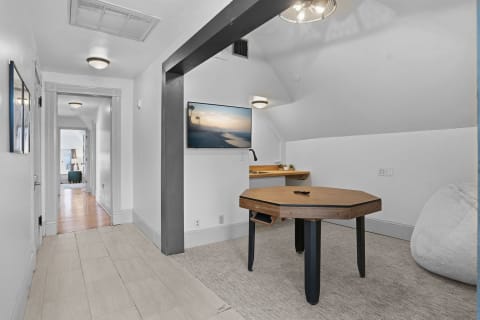 A game room showcasing a poker table, a TV displaying a beach scene, and a cozy bean bag, featuring vaulted ceilings with exposed beams.