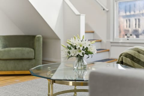 A cozy living space with a glass coffee table and white lilies in a vase, featuring a green armchair and a staircase in the background.