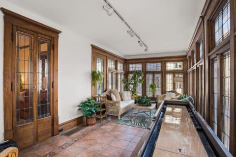 A cozy sunroom featuring rich wooden details, large windows, and plants, complemented by comfortable seating.