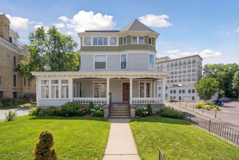 Victorian house with turret and porch, surrounded by a lawn and modern buildings.