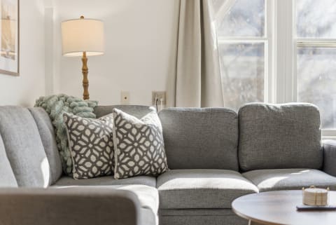 Cozy living room corner with gray sectional sofa, patterned pillows, and a decorative wooden table, bathed in natural light.