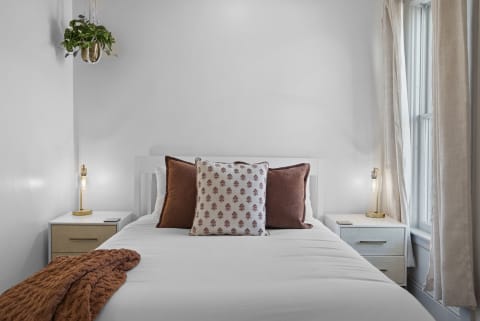 A cozy bedroom with a white bed, earthy pillows, a warm throw, and natural light from the window.